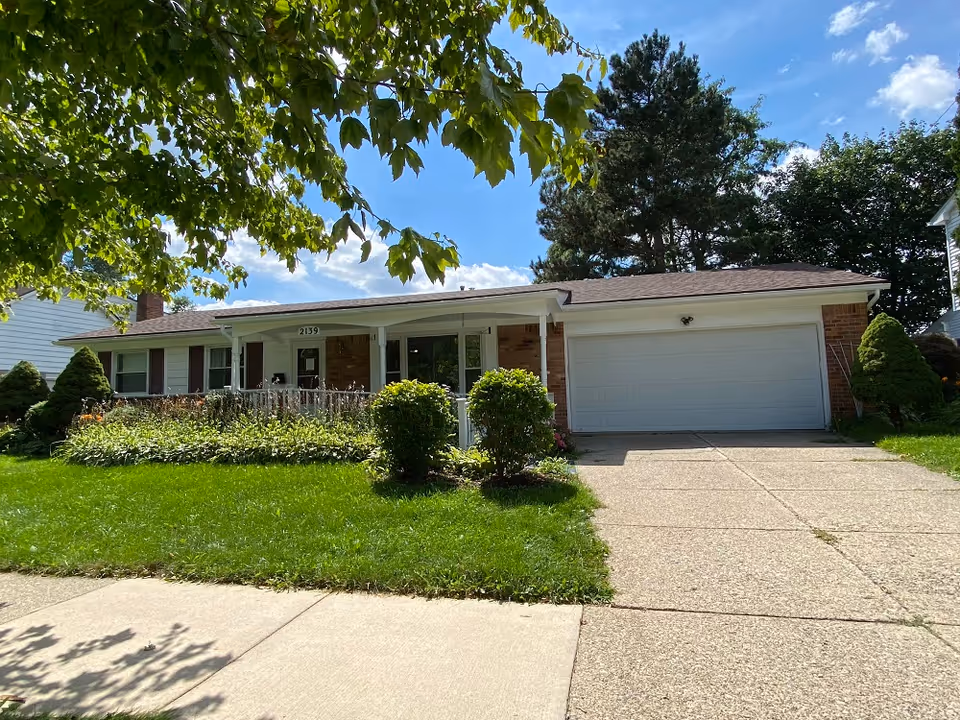 Single-story brick house with a front porch, two bushes in the front yard, a concrete driveway leading to a two-car garage, and a tree with green leaves partially shading the view under a blue sky with some clouds.
