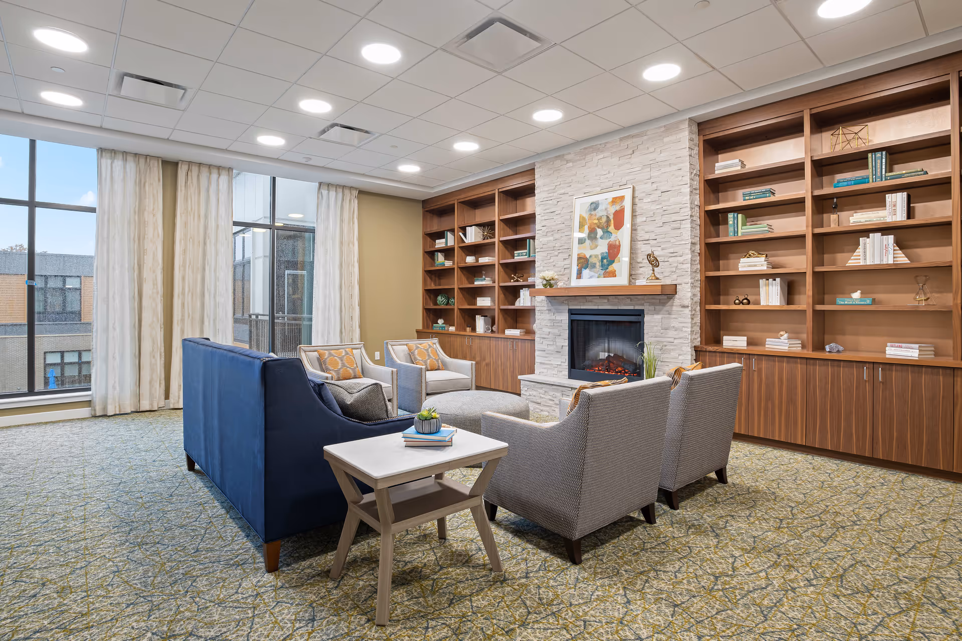 A cozy living room area with a blue sofa, four gray armchairs, a small side table with books and a plant, a stone fireplace with a colorful abstract painting above it, and wooden built-in bookshelves filled with books and decorative items. Large windows with sheer curtains let in natural light.