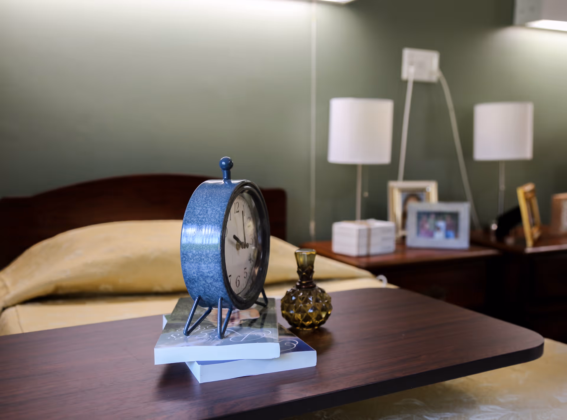 A bedside table with a blue vintage alarm clock, two stacked books, and a small decorative glass bottle. In the background, there is a bed with a beige blanket and two nightstands each with a white lamp and framed photos against a green wall.