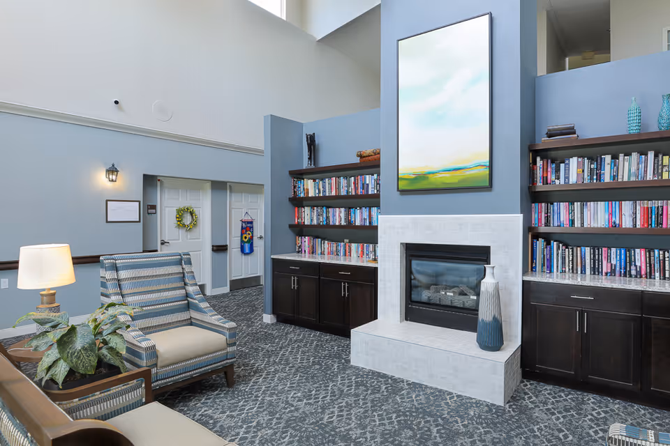 A cozy living room area in an assisted living facility featuring a modern fireplace with a large abstract painting above it. On either side of the fireplace are built-in dark wood bookshelves filled with books and decorative items. There are comfortable striped armchairs and a side table with a lamp and a potted plant. The room has high ceilings and light blue walls.