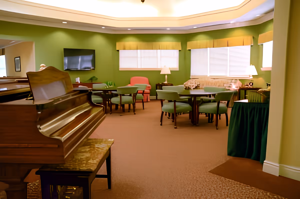 A senior living common room with a piano in the foreground, round tables and chairs, lamps, and windows along a green wall.