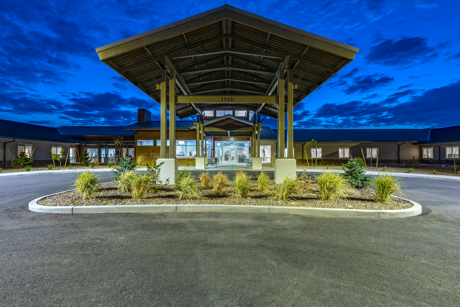 Front exterior view of The Landings Of Prescott Valley facility at dusk, featuring a covered entrance with a peaked roof, landscaped plants, and illuminated windows.