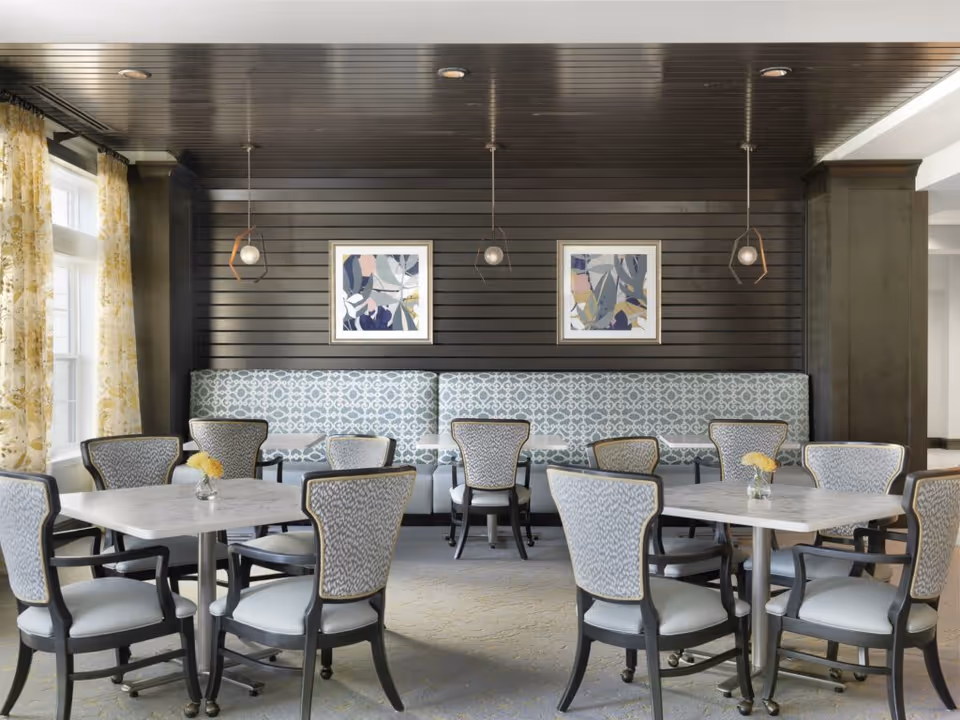 A bright and elegant dining area with multiple square tables surrounded by upholstered chairs. The back wall features dark wood paneling with two framed abstract art pieces and three modern pendant lights hanging from the ceiling. Large windows with yellow floral curtains allow natural light to fill the space.