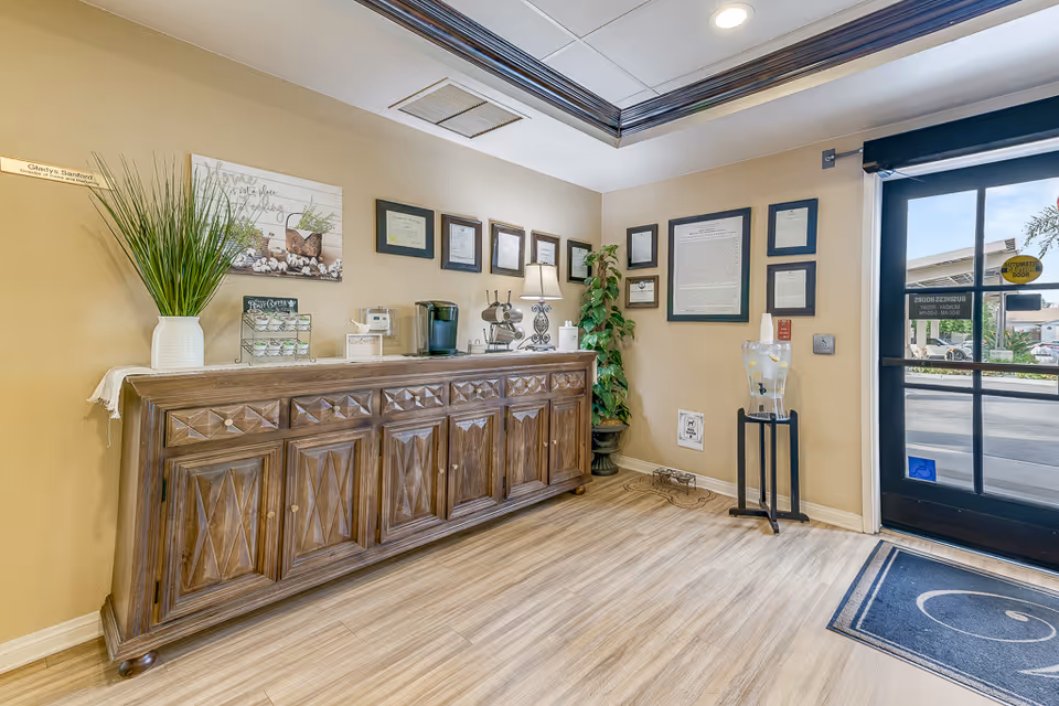 Reception/lobby area with a wooden credenza holding a coffee station and decor, framed certificates on the wall, a water dispenser, and a glass entrance door.