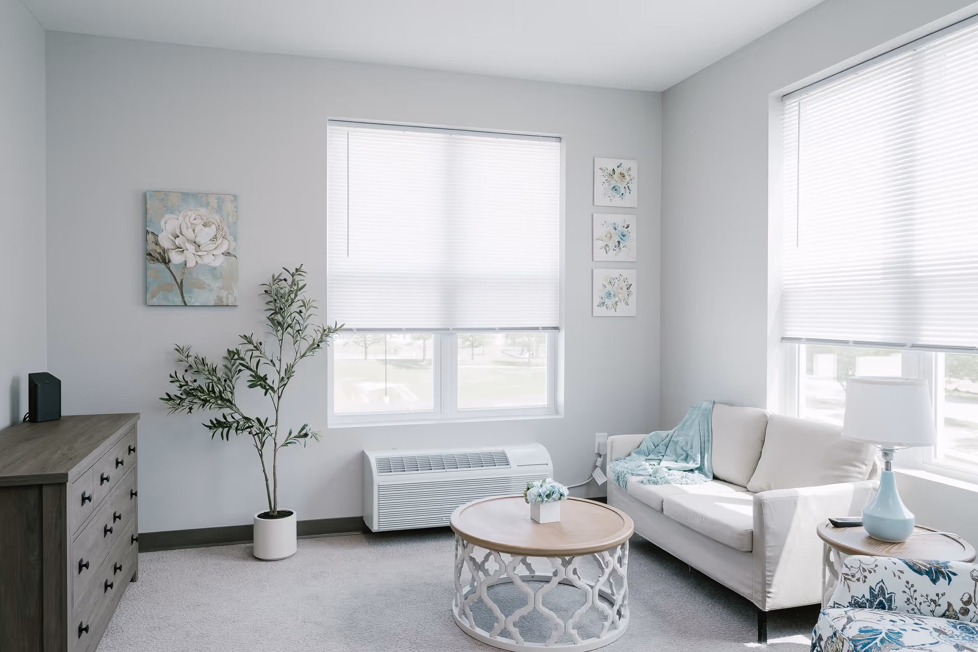 A bright and cozy living room with light gray walls and carpet. The room features a beige sofa with a teal throw blanket, a round wooden coffee table with a small flower arrangement, a floral patterned armchair, a wooden dresser, a potted plant, and two windows with white blinds letting in natural light. There are floral paintings on the walls and a table lamp on a side table next to the sofa.