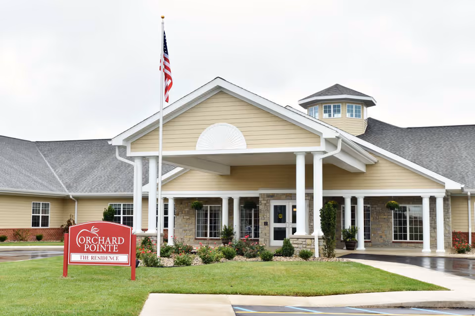 Exterior front view of Orchard Pointe Health Campus building with a covered entrance supported by white columns, an American flag on a flagpole, and a red sign reading 'Orchard Pointe The Residence' on a green lawn.