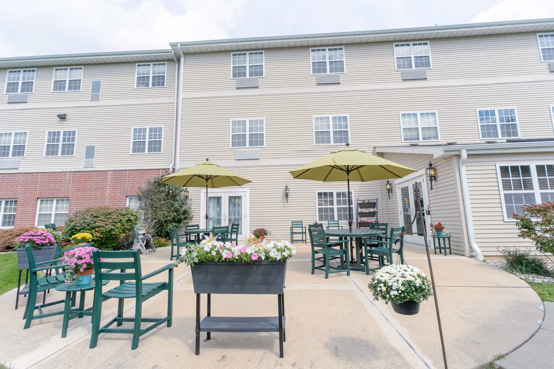 Outdoor patio area at Heritage Woods of Rockford with green chairs and tables under green umbrellas, surrounded by various flower pots and planters. The building exterior is beige with multiple windows and a brick lower section.