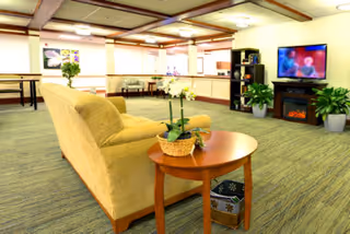 A spacious living room area in an assisted living center featuring a beige couch, a wooden side table with a potted plant, a flat-screen TV mounted on the wall above an electric fireplace, and additional seating and tables in the background. The room has carpeted floors and soft lighting.