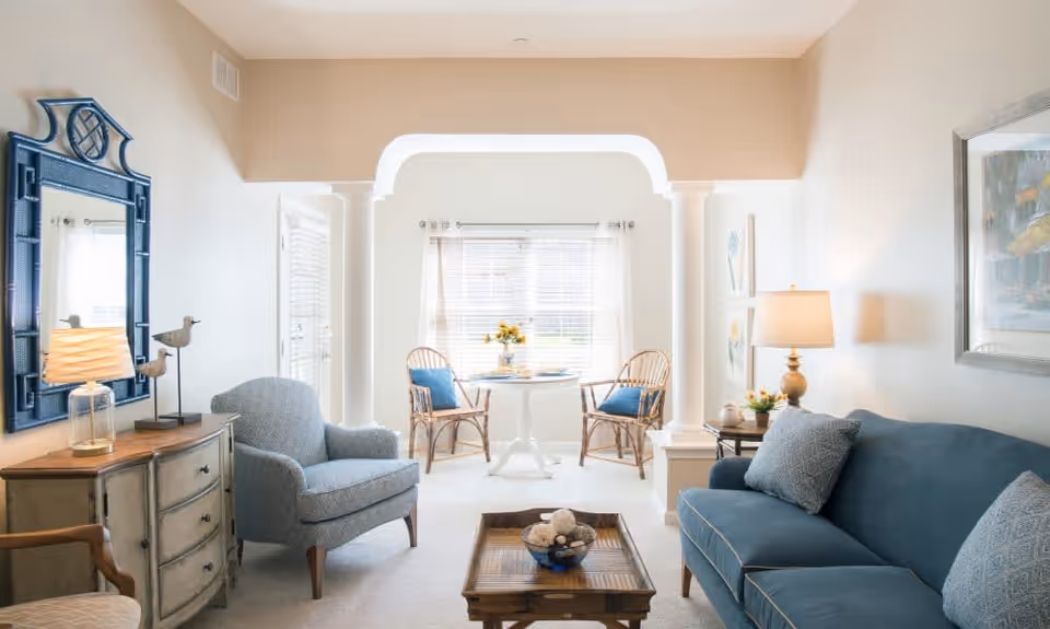 A bright and cozy living room with a blue upholstered sofa and armchair, a wooden coffee table with decorative items, a side table with a lamp and bird figurines, a large mirror on the wall, and a small dining area with two wicker chairs and a round table near a window with sheer curtains.