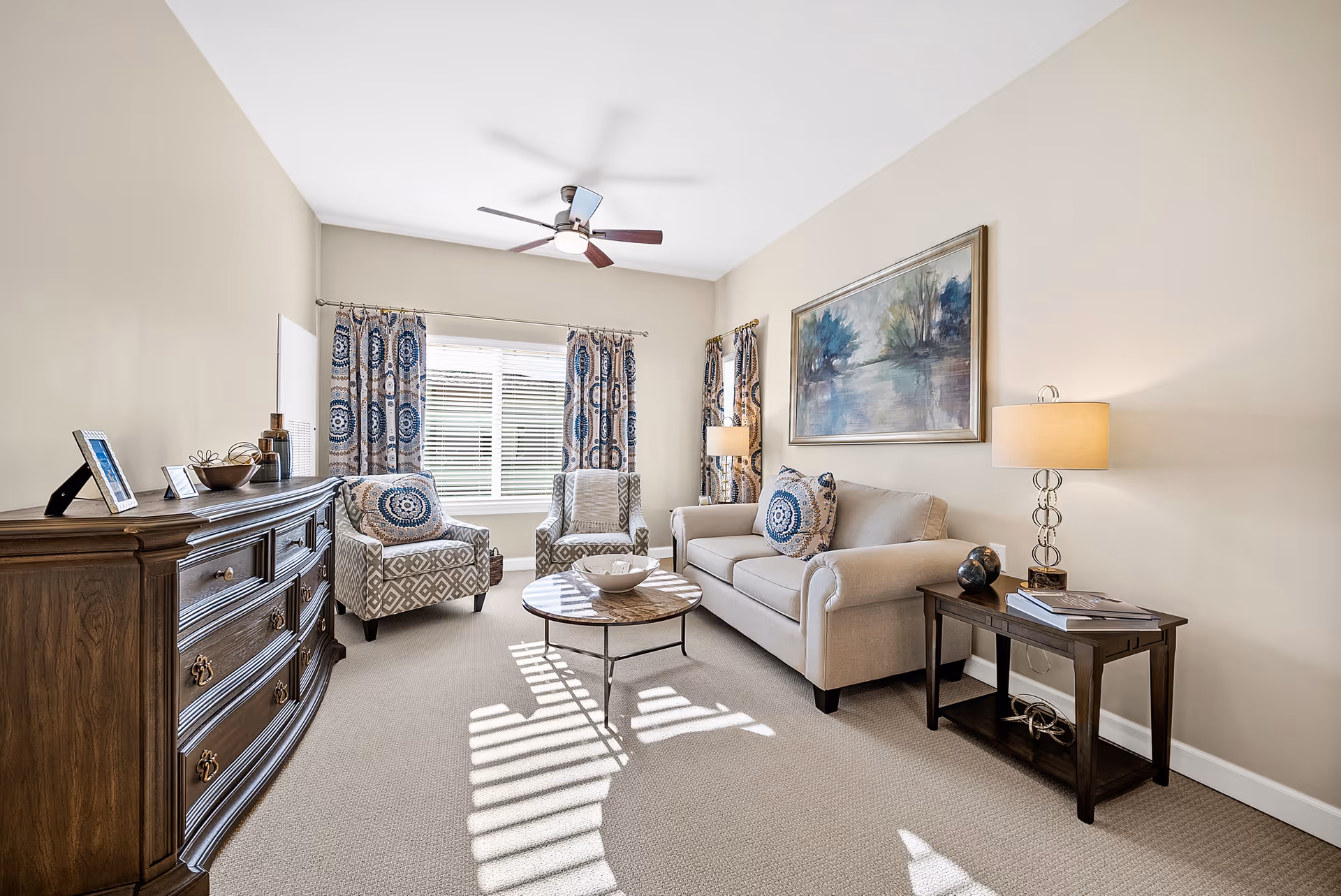 A bright and cozy living room with beige walls and carpet. The room features a beige sofa with patterned blue and white pillows, two matching armchairs with geometric designs, a round coffee table with a bowl on it, a wooden side table with a lamp and books, and a wooden dresser with decorative items. A large window with patterned curtains allows natural light to fill the room, and a ceiling fan is mounted on the white ceiling. A framed landscape painting hangs above the sofa.