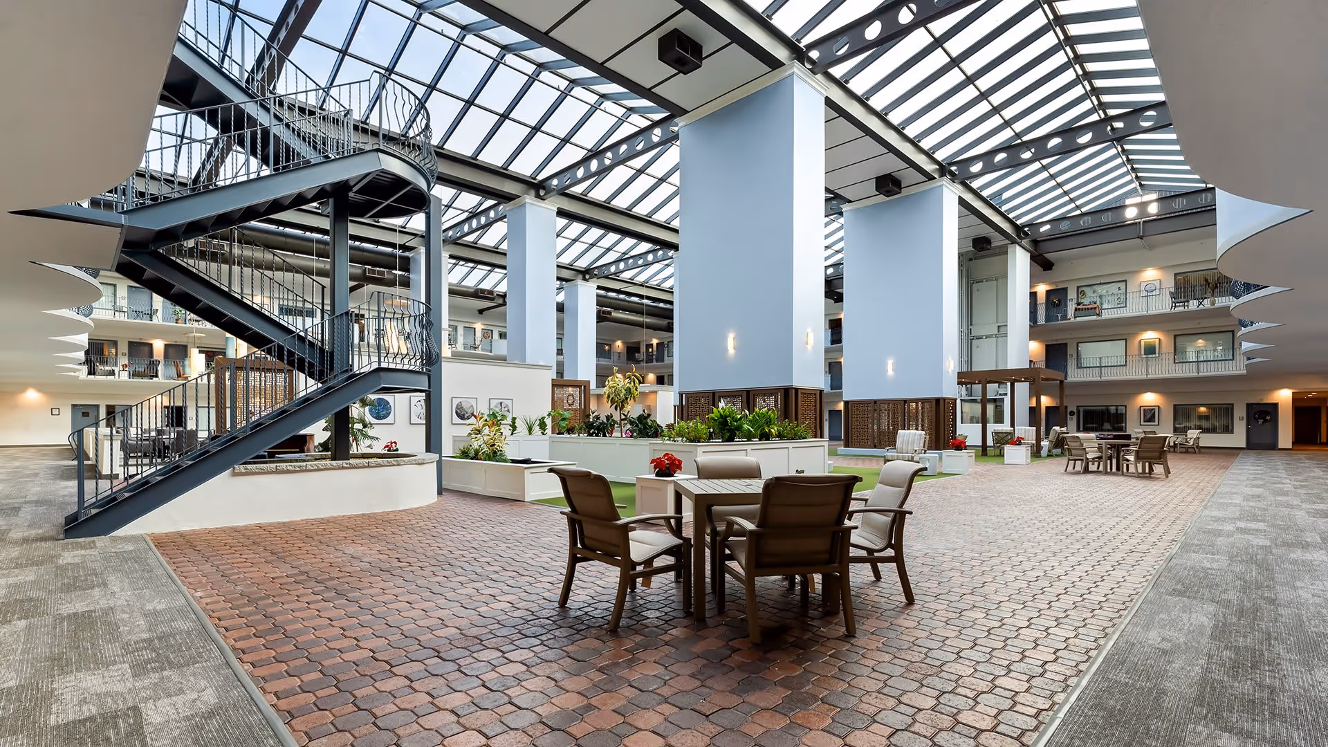 Spacious indoor common area with a high glass ceiling allowing natural light to fill the space. The area features a spiral staircase on the left, several seating arrangements with tables and chairs, and large white pillars. There are plants and decorative elements around the seating areas, and multiple floors with balconies overlooking the space.