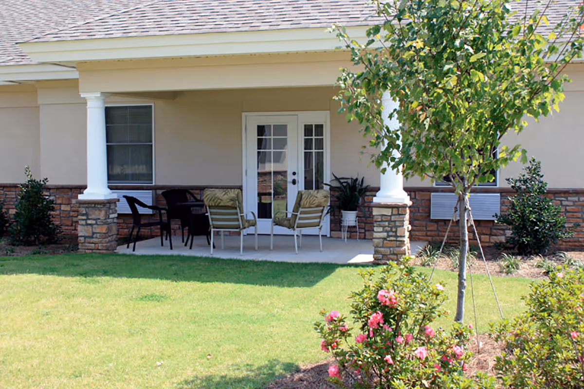 Covered patio with chairs and columns in front of a residential building, with a lawn and flowering shrubs.