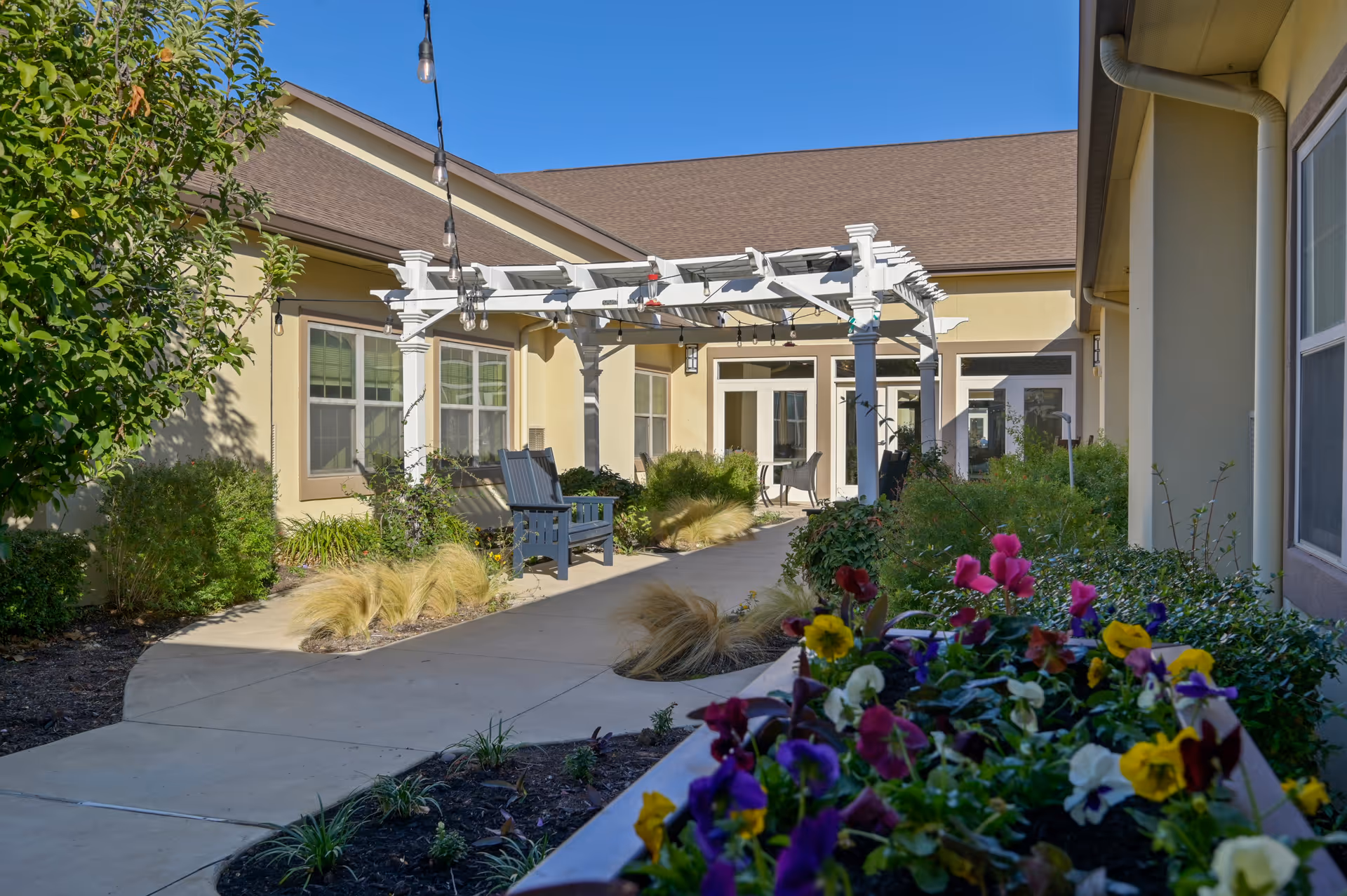 Sunny courtyard at a senior living facility featuring a white pergola over a paved walkway, benches, and colorful flowerbeds.
