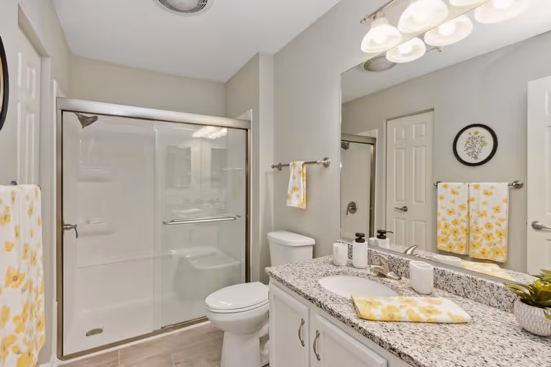 A clean and modern bathroom featuring a glass-enclosed shower with sliding doors, a white toilet, and a granite countertop with a sink. Yellow floral towels hang on towel racks, and a small potted plant is placed on the counter. A large mirror is mounted above the sink, reflecting the opposite wall with a door and framed floral artwork.