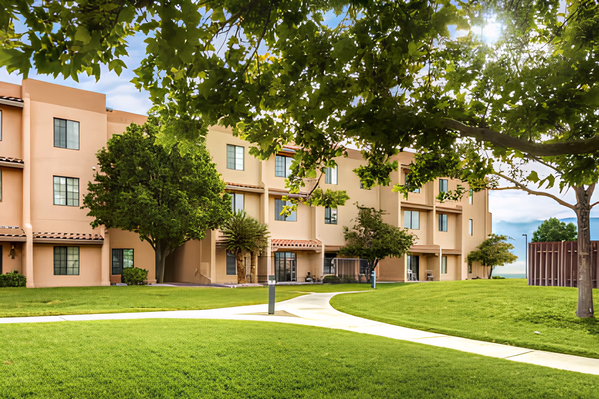 Three-story beige senior living building with a green lawn, trees, and a curving sidewalk under leafy branches.
