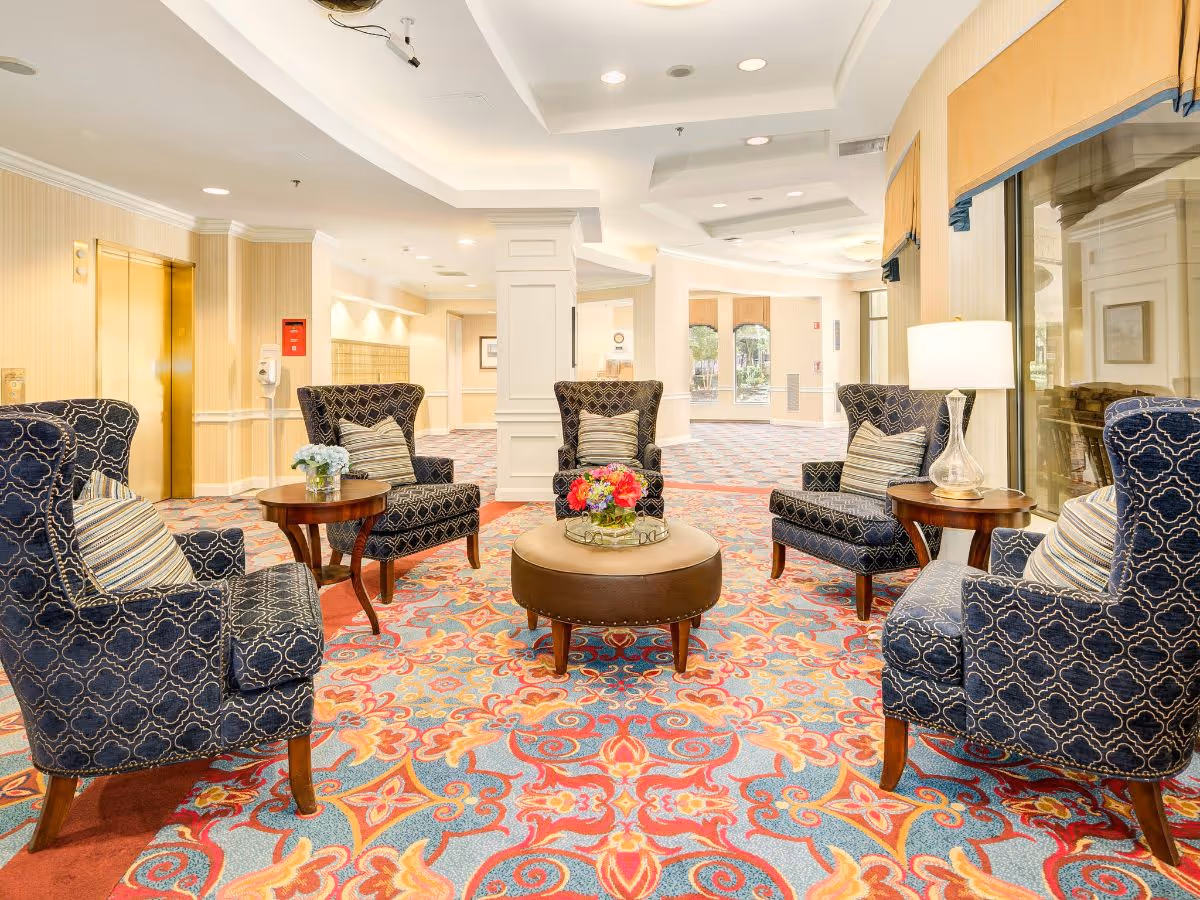 A bright and spacious seating area in a senior living facility featuring four patterned armchairs arranged around a round ottoman with a flower arrangement on top. There are two small wooden side tables with a lamp and a small flower vase. The room has a colorful patterned carpet, large windows with yellow valances, and a gold elevator door in the background.