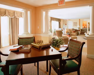 Sunlit communal lounge with upholstered chairs, columns, and a table holding a chessboard and books.