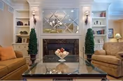 A cozy living room with two beige sofas facing each other, a glass coffee table with a decorative bowl in the center, a fireplace with a mirrored wall above it, and built-in shelves on either side holding books and decorative items. Two tall potted plants flank the fireplace.