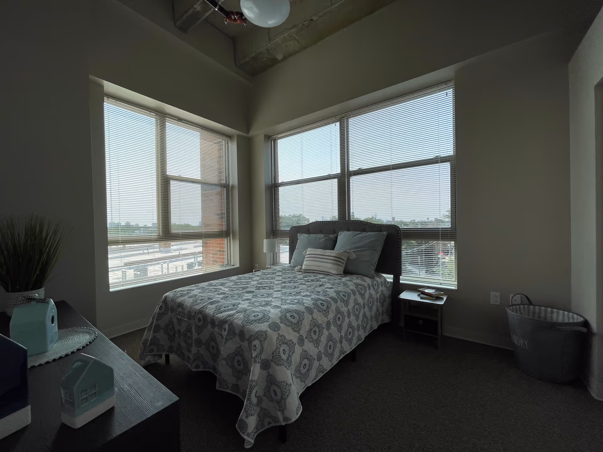 A bedroom with a bed covered in a patterned bedspread and several pillows, positioned near large windows with blinds partially open, allowing natural light to enter. There is a small nightstand with a lamp and a book, a laundry basket, and a dresser with decorative items including a small house-shaped ornament and a plant.