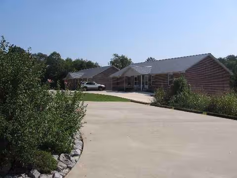 Single-story lodge-style senior living building with a driveway, greenery, and parked cars.