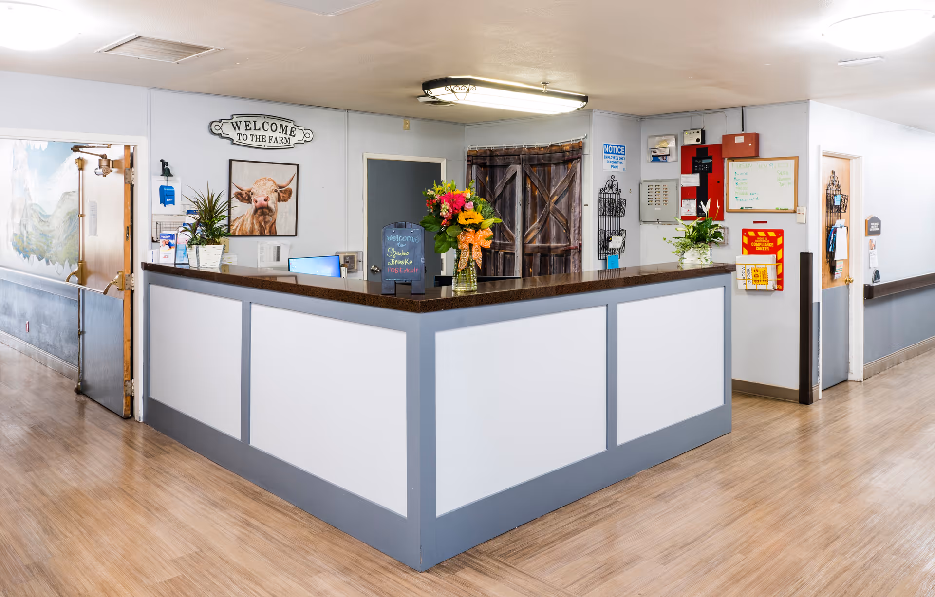 Reception desk area in a senior living facility with a 'Welcome to the Farm' sign and a picture of a cow on the wall behind the desk. The desk has a dark countertop with flowers and a small chalkboard sign. The hallway has light-colored walls and wood flooring, with doors and safety equipment visible.