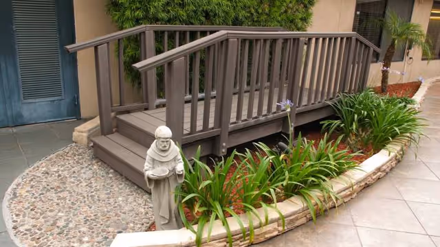 Wooden ramp with handrails leading to a doorway beside a curved planted bed and a small stone statue in an outdoor courtyard.