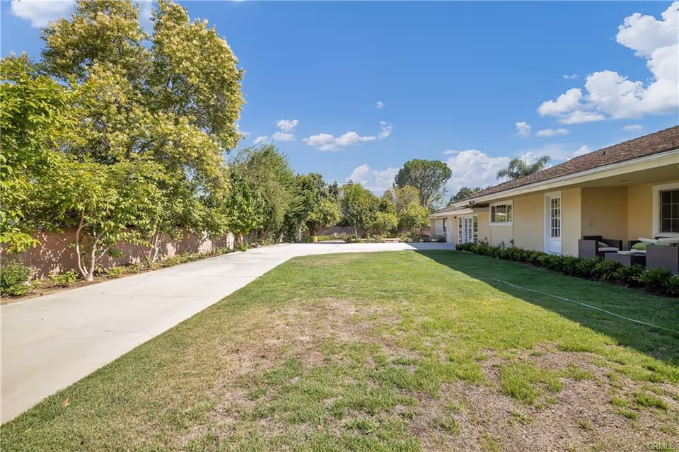 Outdoor view of a senior living facility showing a large grassy lawn with some patches of dry grass, a concrete pathway on the left, and a single-story building with a covered patio area on the right. Trees and shrubs line the left side of the pathway under a blue sky with scattered clouds.