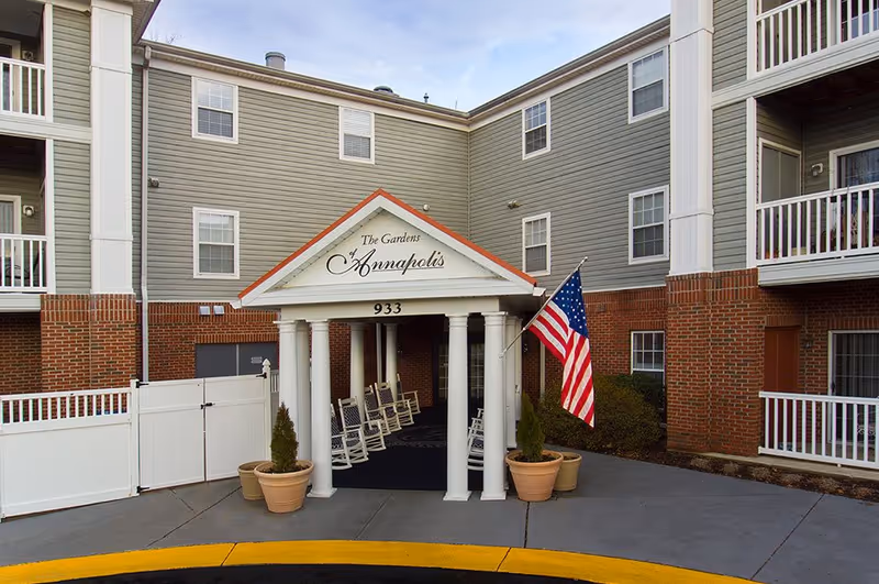 Entrance canopy of The Gardens of Annapolis senior living building with rocking chairs and an American flag.