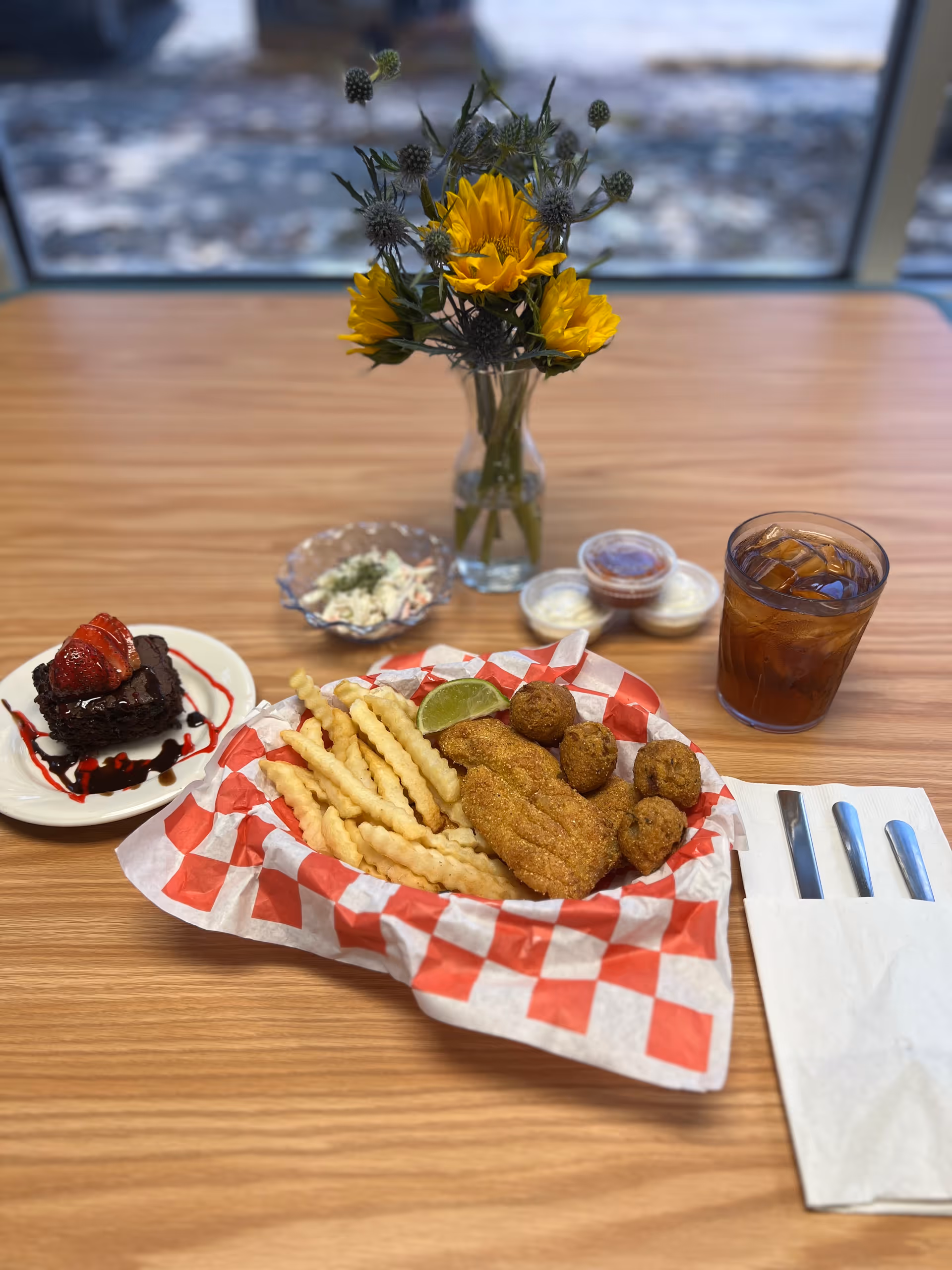 Tray of fried food and crinkle-cut fries with iced tea, dessert, coleslaw, and a vase of yellow flowers on a wooden dining table by a window.