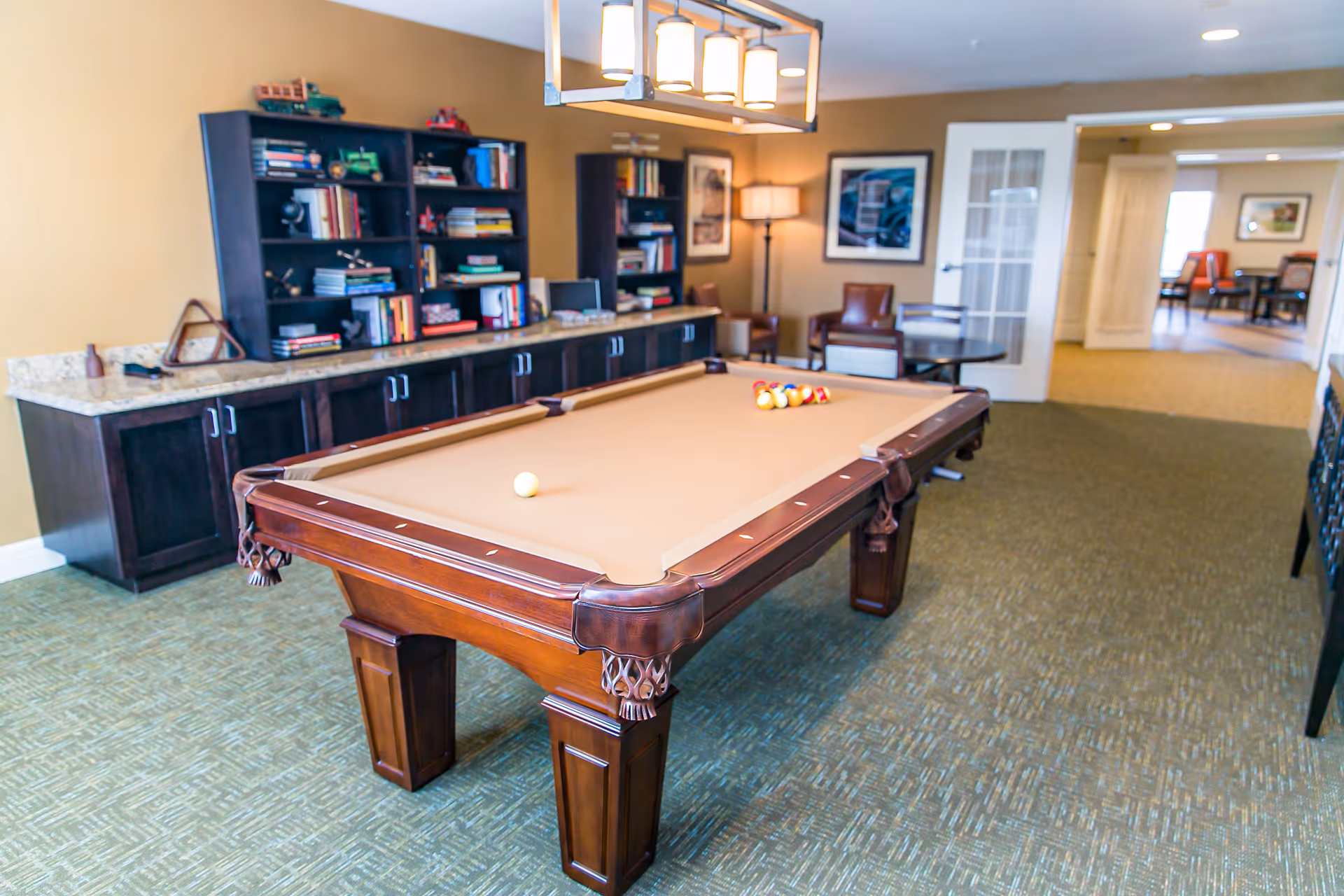 A well-lit game room featuring a brown pool table with billiard balls arranged on it. Behind the pool table is a dark wooden cabinet with shelves filled with books and decorative items. The room has beige walls, carpeted flooring, and a modern light fixture hanging above the pool table. In the background, there are chairs, a floor lamp, and framed artwork on the walls.