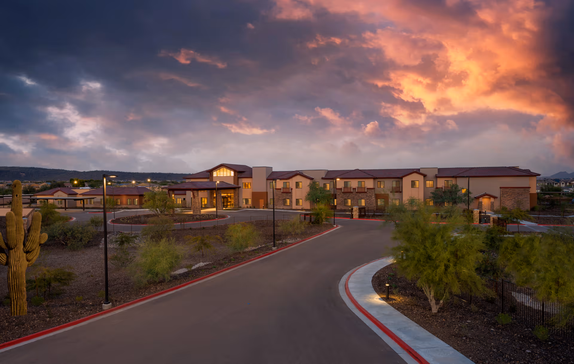 Exterior front view of an assisted living facility at dusk with a curved driveway, desert landscaping, and dramatic colorful clouds.