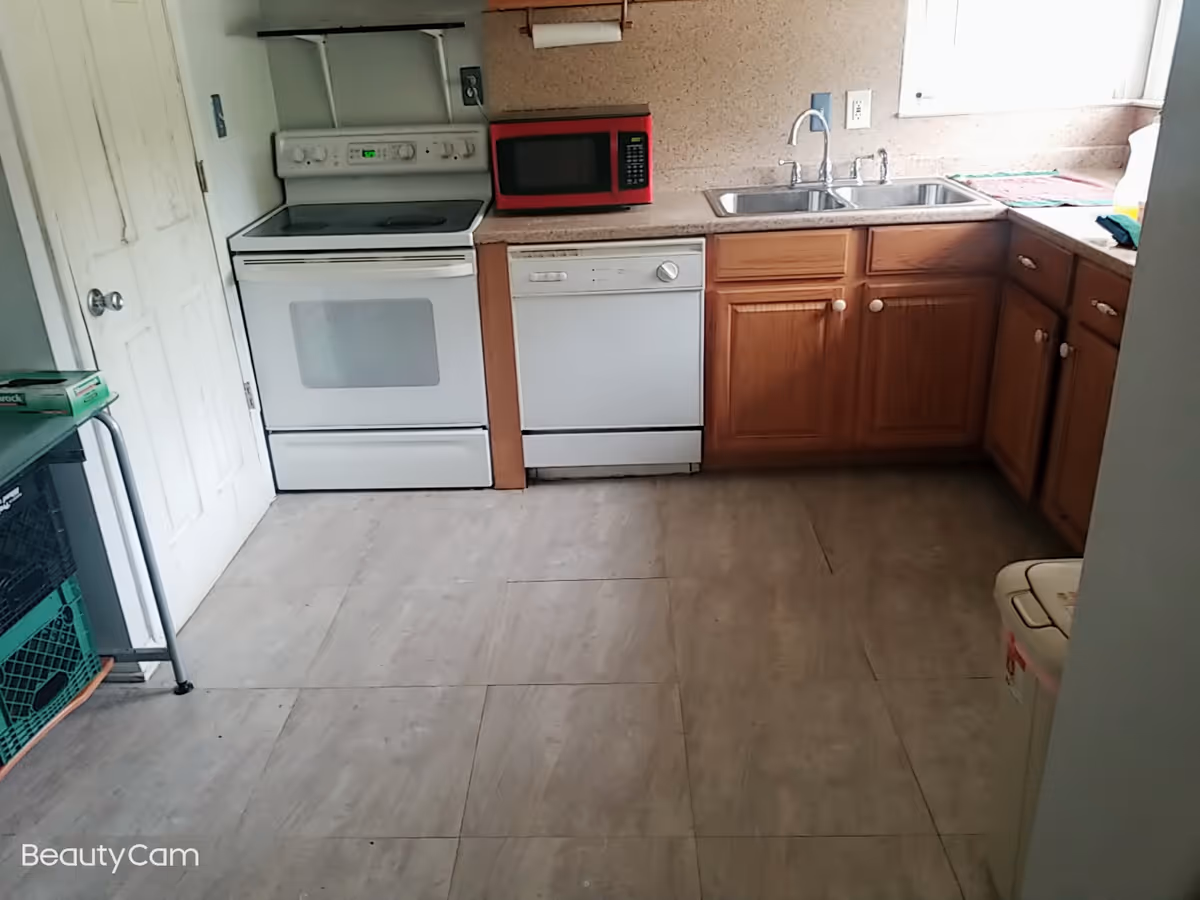 A kitchen with a white electric stove, a red microwave, a dishwasher, wooden cabinets, a double sink, and a window above the sink. The floor is tiled and there is a door on the left side. A trash can is partially visible on the right.