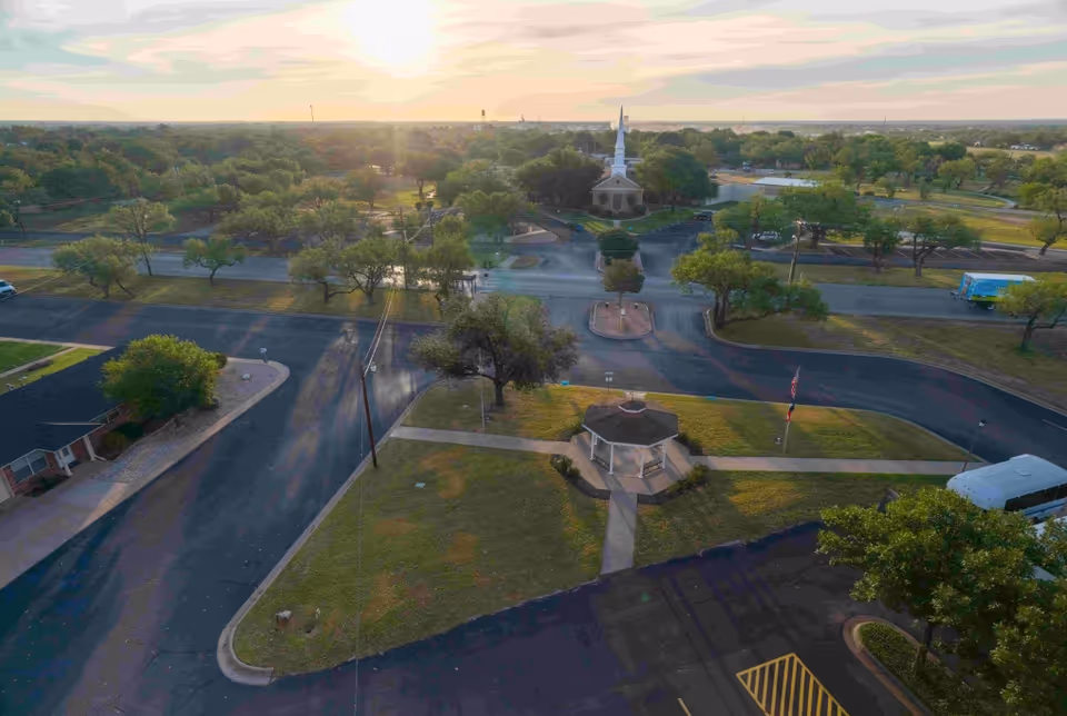 Aerial view of a retirement community with a small gazebo in the foreground, a church with a tall steeple in the background, surrounded by trees and roads under a setting sun.