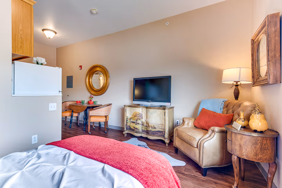 A cozy senior living room with a beige armchair adorned with a red pillow and blue throw blanket, a wooden side table with decorative items and a lamp, a TV on a painted cabinet, a small dining table with two chairs, and part of a bed with a red blanket in the foreground.