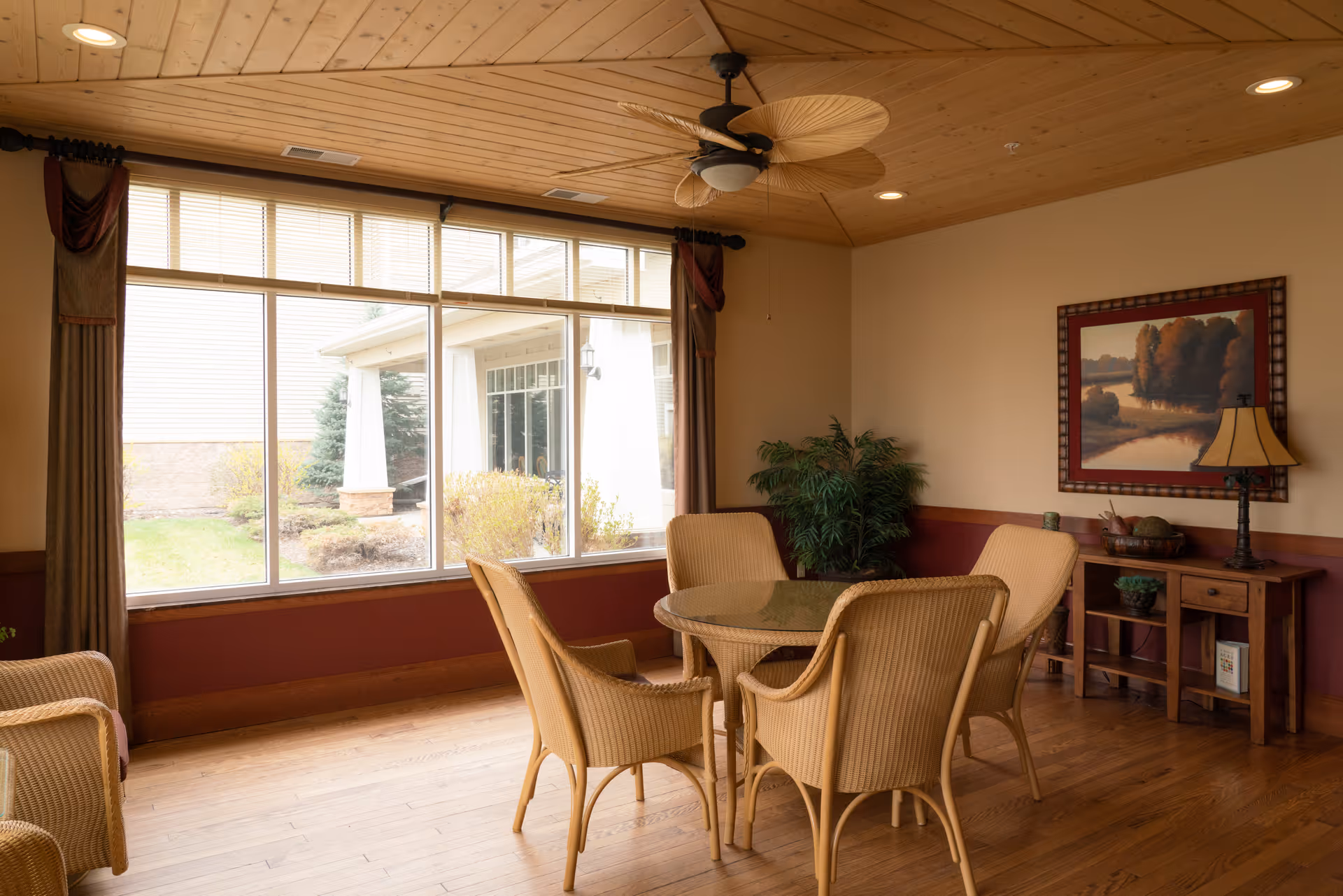 Sunlit common room with a round glass-top table surrounded by wicker chairs, a large window, wooden floors and a ceiling fan.