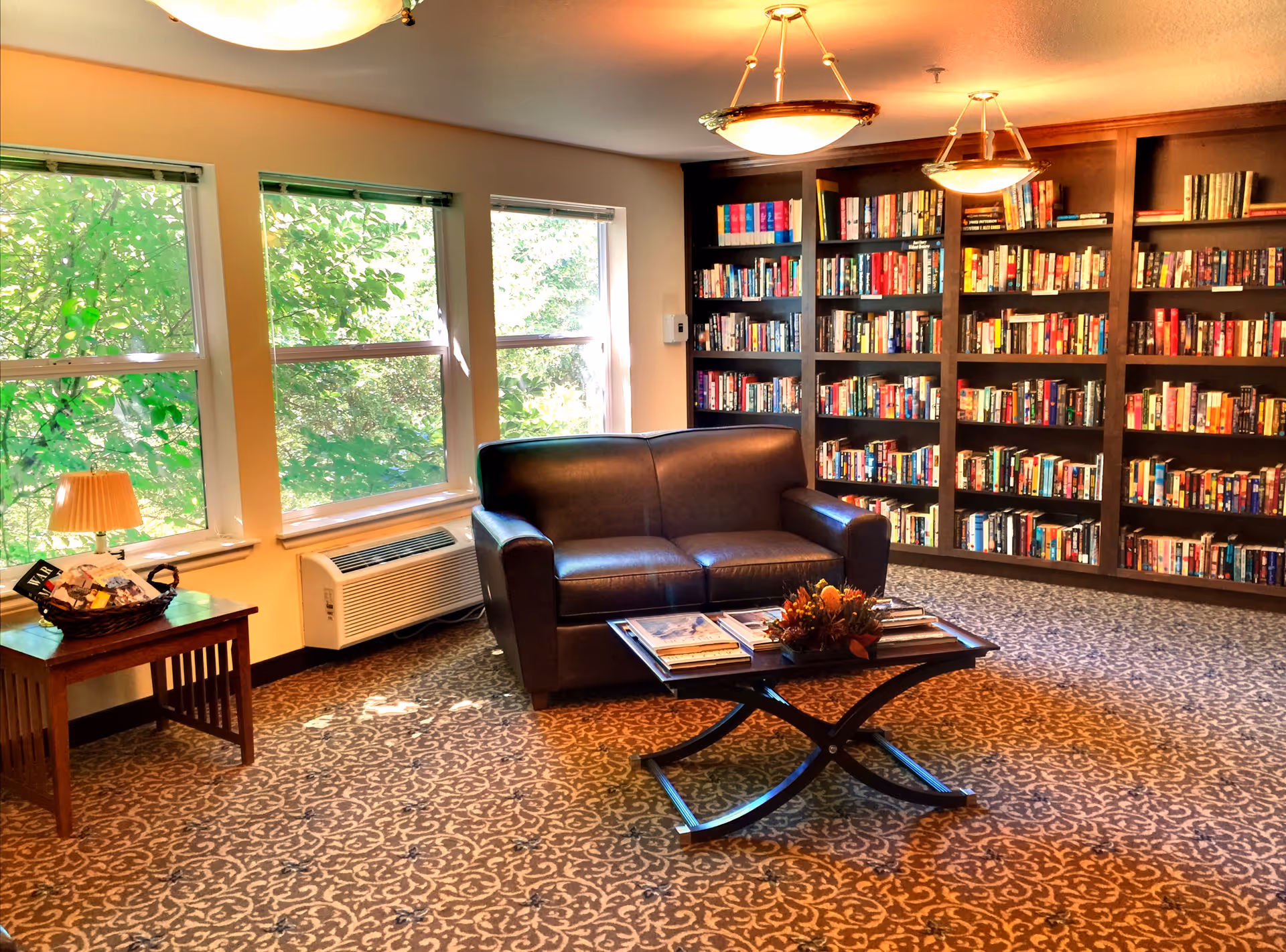 A cozy library-style sitting area with a leather loveseat, coffee table, and a wall of bookshelves beside windows.