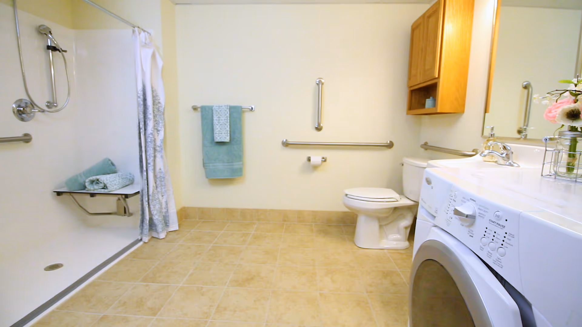 A spacious bathroom with beige tiled floor, a walk-in shower with a handheld showerhead and a white curtain, a white toilet with grab bars on the walls, a white sink with a chrome faucet, a wooden cabinet above the sink, a washing machine, and teal towels hanging on a towel rack and folded on a shower bench.