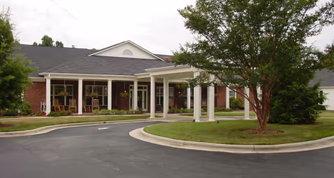 Front exterior view of a single-story brick building with a covered entrance supported by white columns, surrounded by a circular driveway and landscaped with green grass and trees.