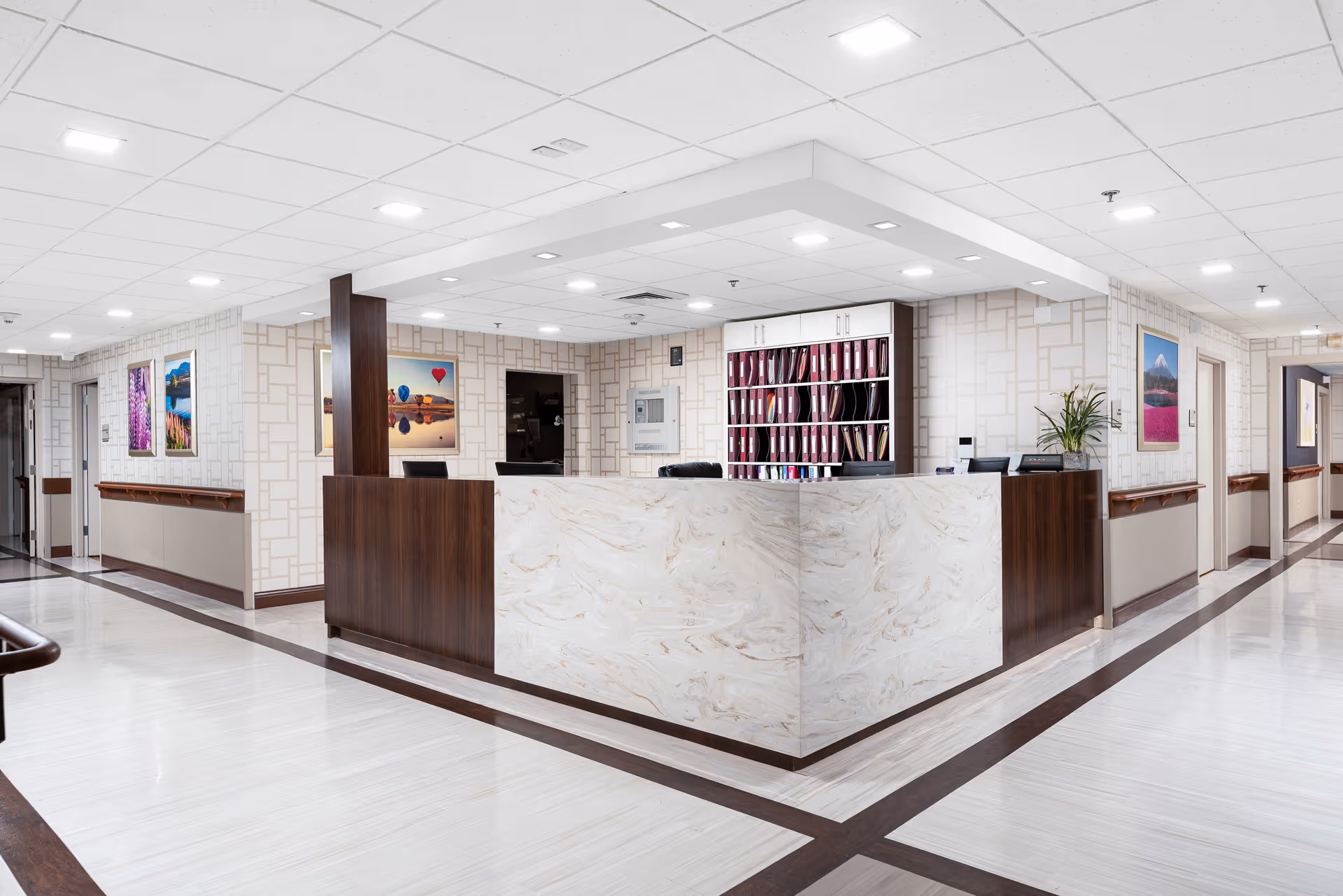 A clean and modern reception desk area in a senior living facility with a marble-patterned front counter and dark wood accents. Behind the desk are shelves filled with organized files and a computer monitor. The walls are decorated with colorful landscape paintings, and the hallway extends on both sides with handrails along the walls. The ceiling has recessed lighting panels providing bright illumination.