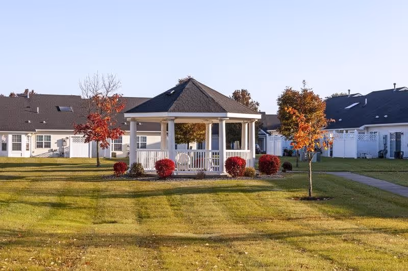 A white gazebo with a dark roof stands in the middle of a well-maintained grassy area with small trees and red bushes. Surrounding the gazebo are single-story residential buildings with white fences and dark roofs under a clear blue sky.