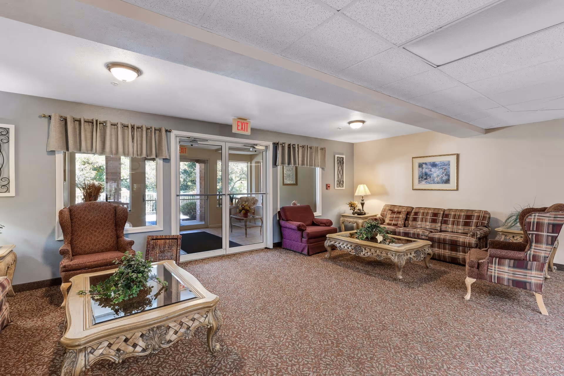 A cozy living room area with patterned carpet, a glass-top coffee table with a plant, several upholstered armchairs and a plaid sofa. There are two windows with beige curtains and a glass door with an exit sign above it. A table lamp and framed artwork decorate the walls.