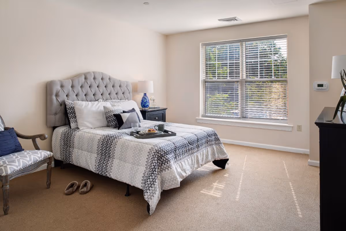 Sunlit bedroom featuring a tufted headboard bed dressed in patterned bedding, a nightstand with lamp, an upholstered chair, and a large window with blinds.