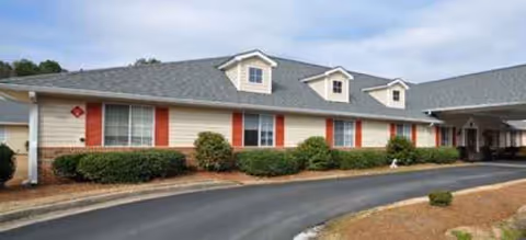 Single story senior living facility exterior with red shutters and manicured shrubs along the driveway.