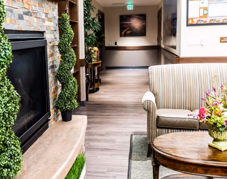 A cozy interior hallway area in a senior living facility featuring a stone fireplace with two spiral-shaped topiary plants on the mantel. To the right, there is a striped upholstered armchair next to a wooden coffee table with a floral arrangement. The hallway has light wood flooring, a small table with flowers further down, and an exit sign above a door at the end.