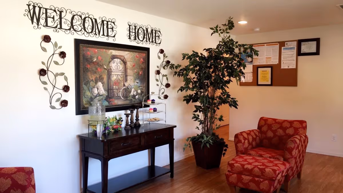 A cozy interior reception area with a 'WELCOME HOME' wall sign, a console table, large potted plant, and a red patterned armchair with ottoman.