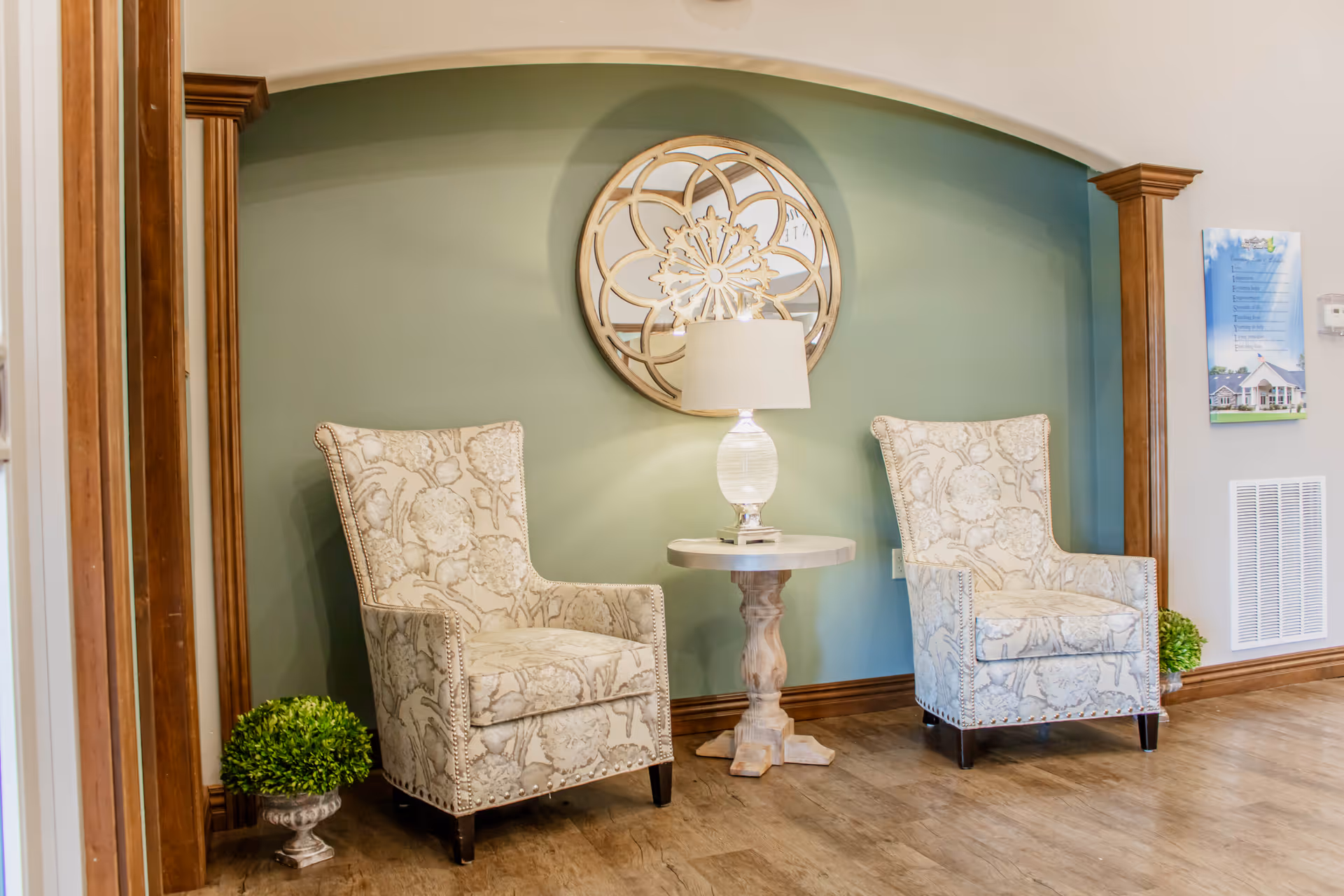 Two patterned armchairs flank a small round table with a lamp beneath a decorative round mirror in a cozy seating alcove.