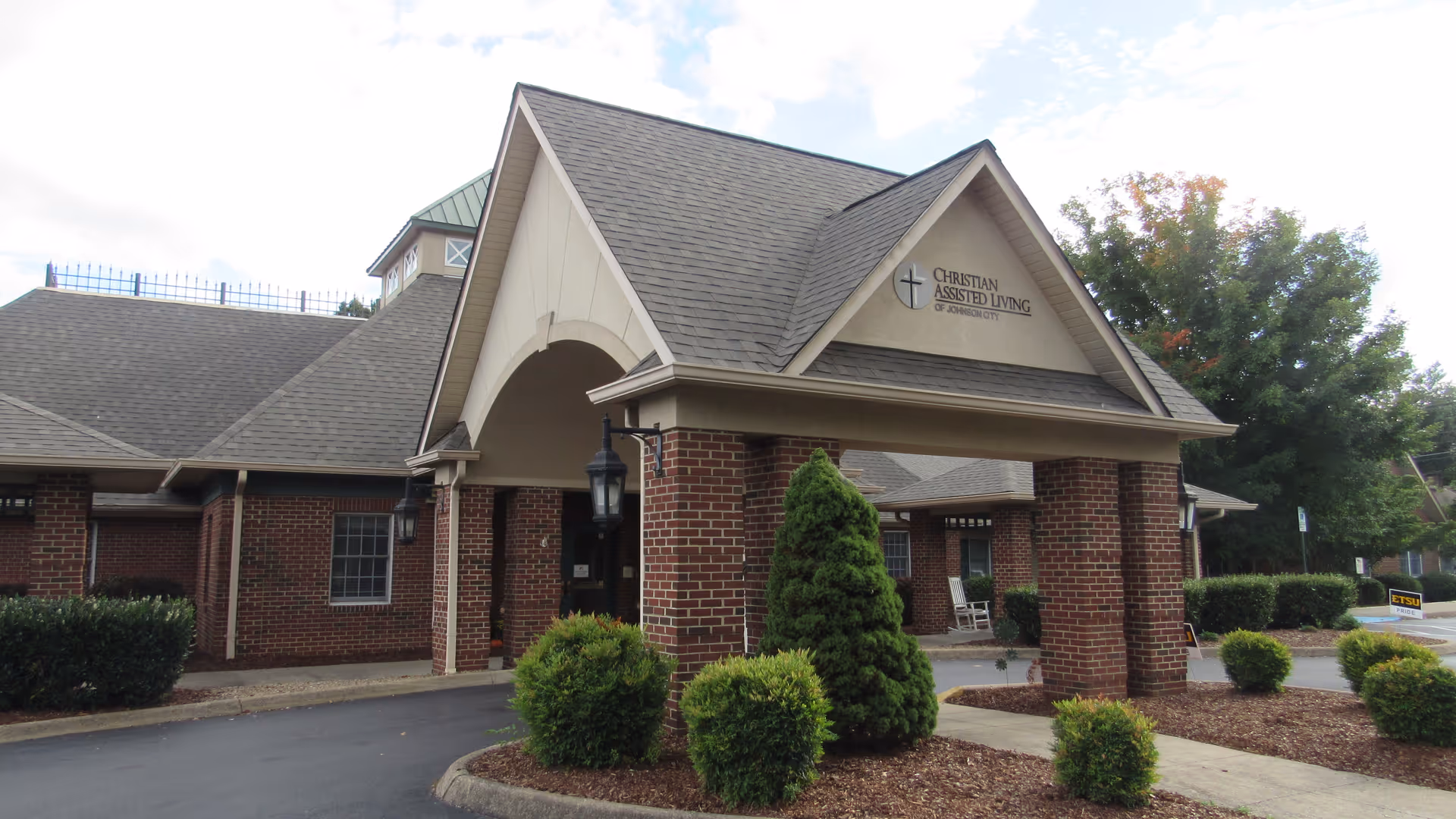 Exterior front entrance of Christian Assisted Living of Johnson City, featuring a brick building with a covered driveway supported by brick columns, surrounded by neatly trimmed bushes and trees.