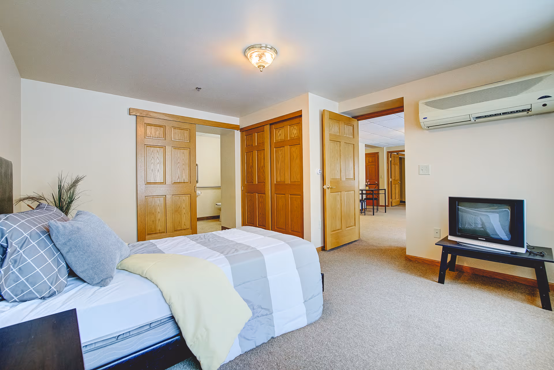 A bedroom in Nature's Point Assisted Living featuring a neatly made bed with gray and white bedding and multiple pillows. The room has beige walls, a ceiling light fixture, wooden doors including a closet and an open door leading to another room. A small TV sits on a black stand beneath a wall-mounted air conditioning unit. The floor is carpeted in a neutral tone.