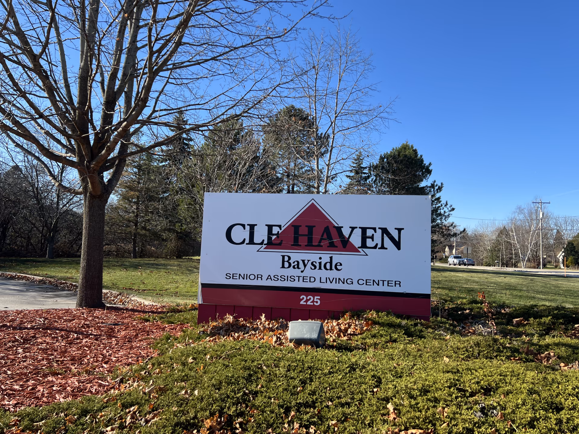 Outdoor view of a sign for CLE HAVEN Bayside Senior Assisted Living Center with the number 225 at the bottom. The sign is surrounded by mulch, grass, and trees under a clear blue sky.
