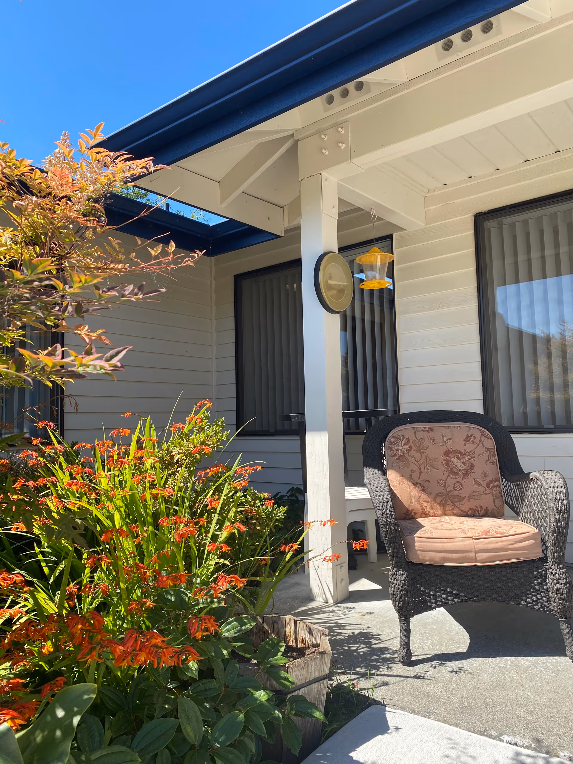 Outdoor patio area at Logan Creek Retirement Community with a wicker chair featuring a floral cushion, a hanging bird feeder, and vibrant orange flowers and green plants under a clear blue sky.